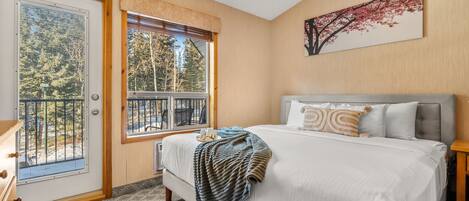 A bedroom with a large bed, a window with a view of trees, and a door with a view of a balcony.