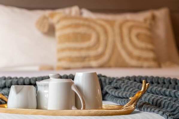 Un plateau de petit-déjeuner soigneusement préparé, avec un pichet, deux tasses et une théière, posé sur un lit agrémenté d’un oreiller à rayures et d’une couverture tricotée.