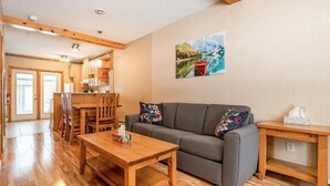 Una sala de estar acogedora con suelo de madera, un sofá gris con cojines de colores, una mesa de centro de madera y un cuadro de un barco en un lago en la pared.