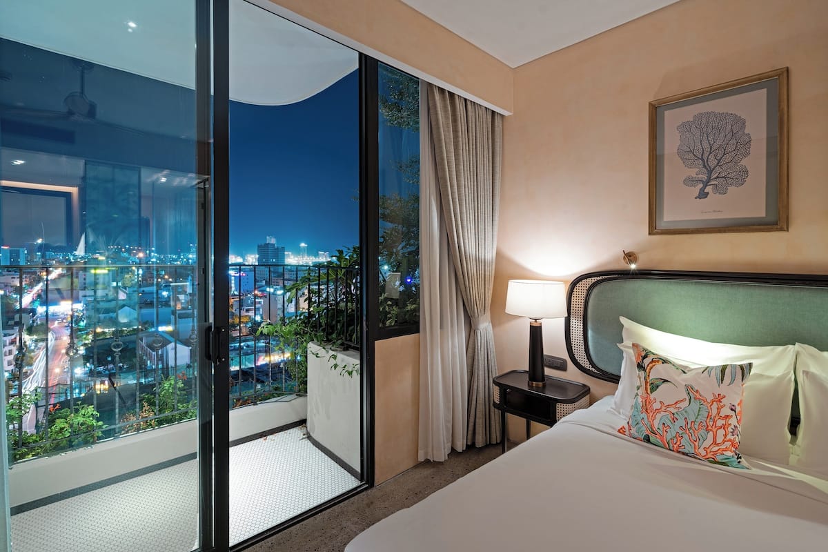 Ein Hotelzimmer mit großem Balkon, einem Bett mit dekorativen Kissen, einem Nachttisch mit Lampe und einem gerahmten Bild an der Wand.