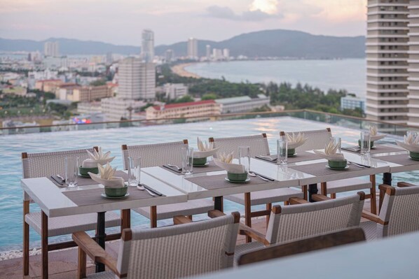 A dining table set with white placemats, green bowls, and clear glasses, overlooking a coastal cityscape. - L'amor Boutique Hotel Quy Nhon (Quy Nhon Nam)