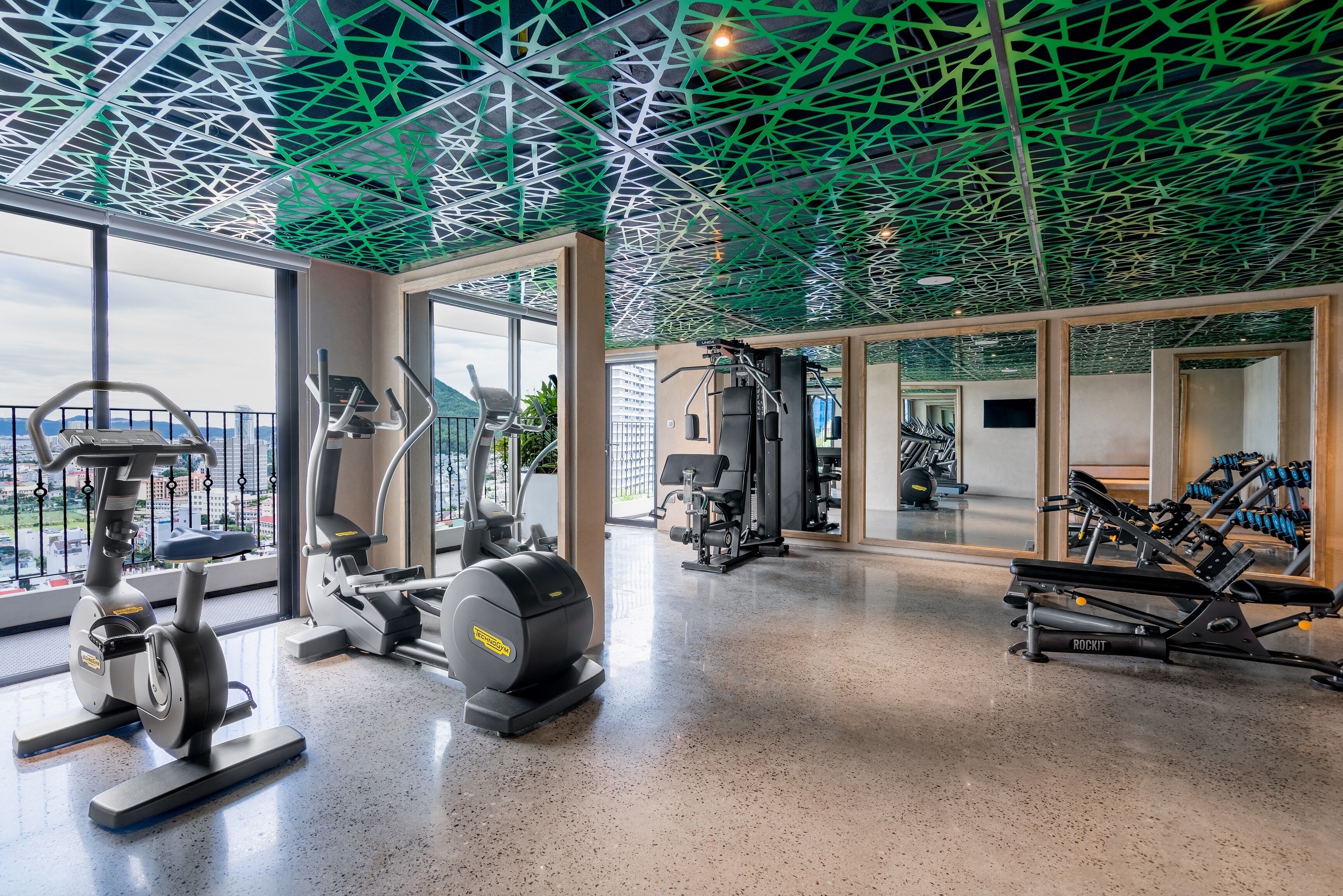 A modern gym with various exercise equipment, large windows, and a unique green ceiling design.