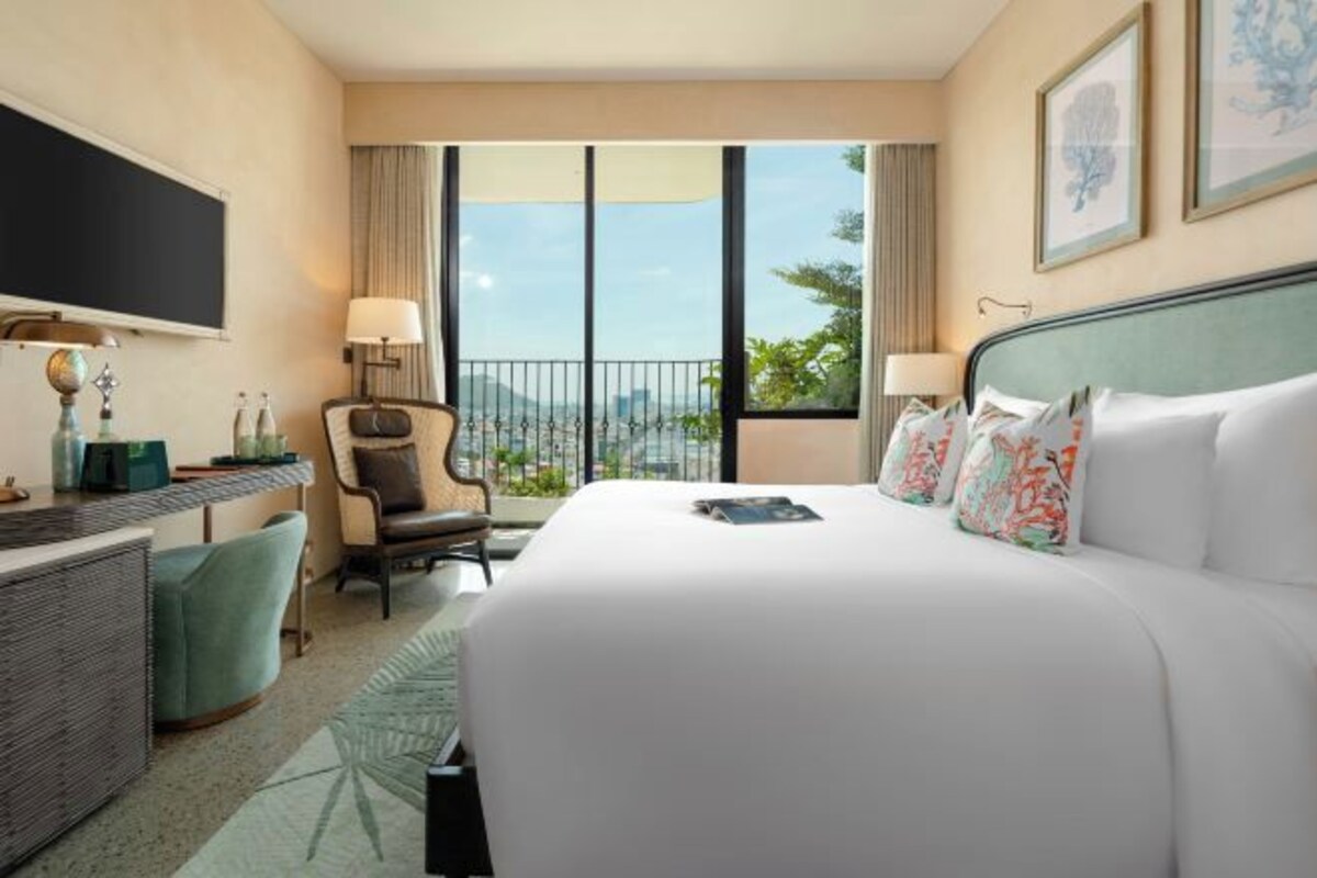 A hotel room with a large bed, a desk, a chair, and a view of the city through large windows.