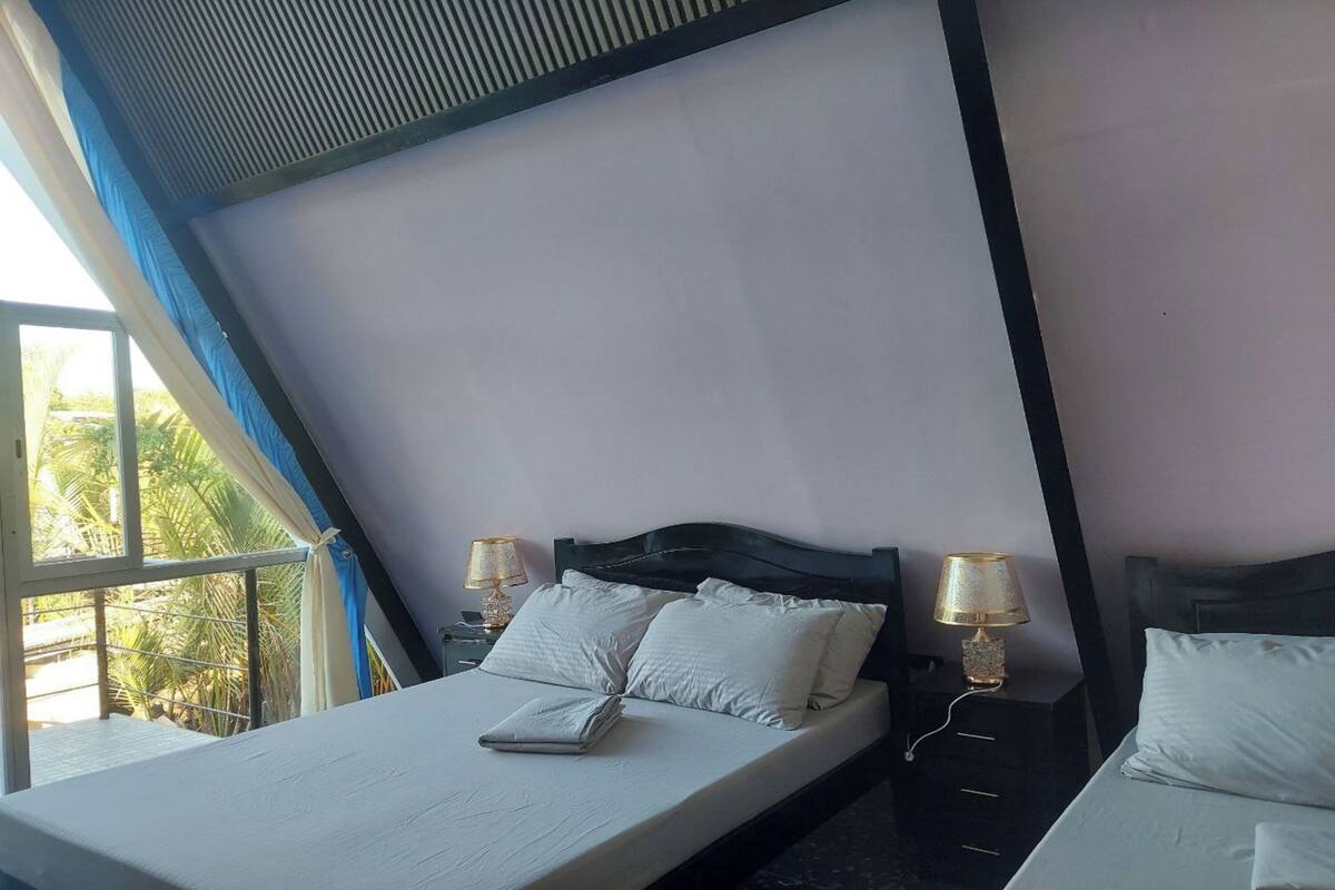 A bedroom with a bed, two bedside tables with lamps, a window with curtains, and a view of greenery outside.