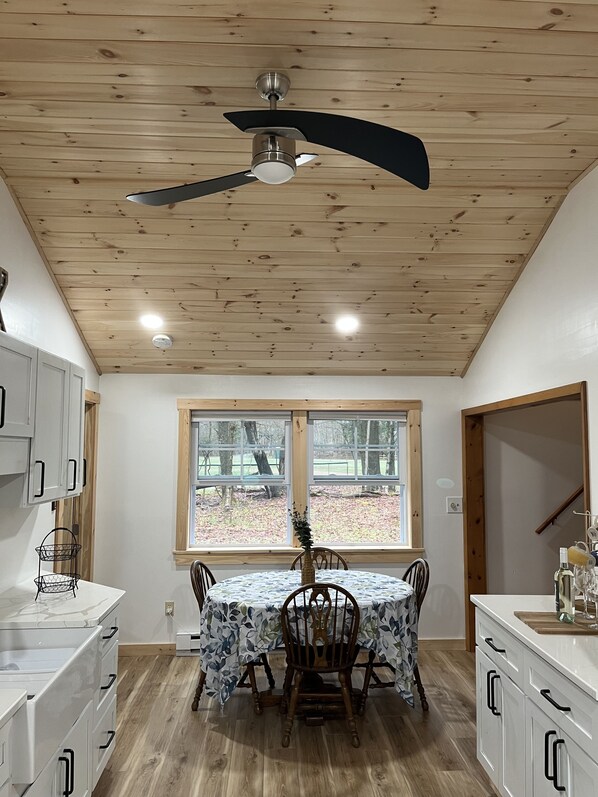 Una cocina con una mesa de comedor, sillas, armarios blancos, un ventilador de techo y una ventana con vista a árboles.