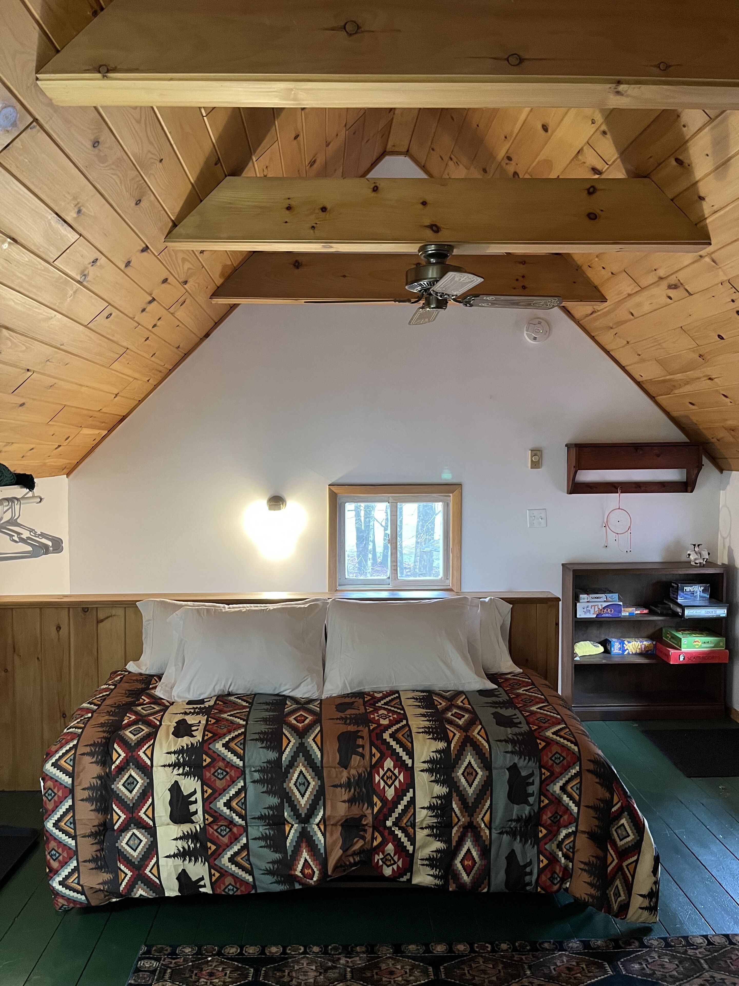 A bedroom with a sloped ceiling, a bed with patterned bedding, a window, and a bookshelf with board games.