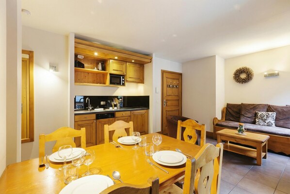 A dining area with a wooden table set for four, a kitchen with wooden cabinets and a microwave, and a brown sofa with a cushion.