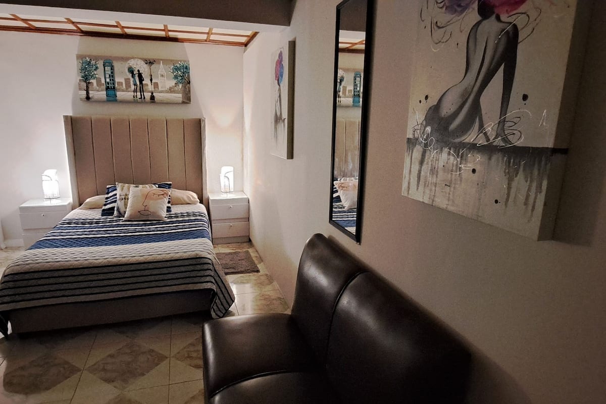 A bedroom with a bed, a chair, a headboard, and artwork on the walls.