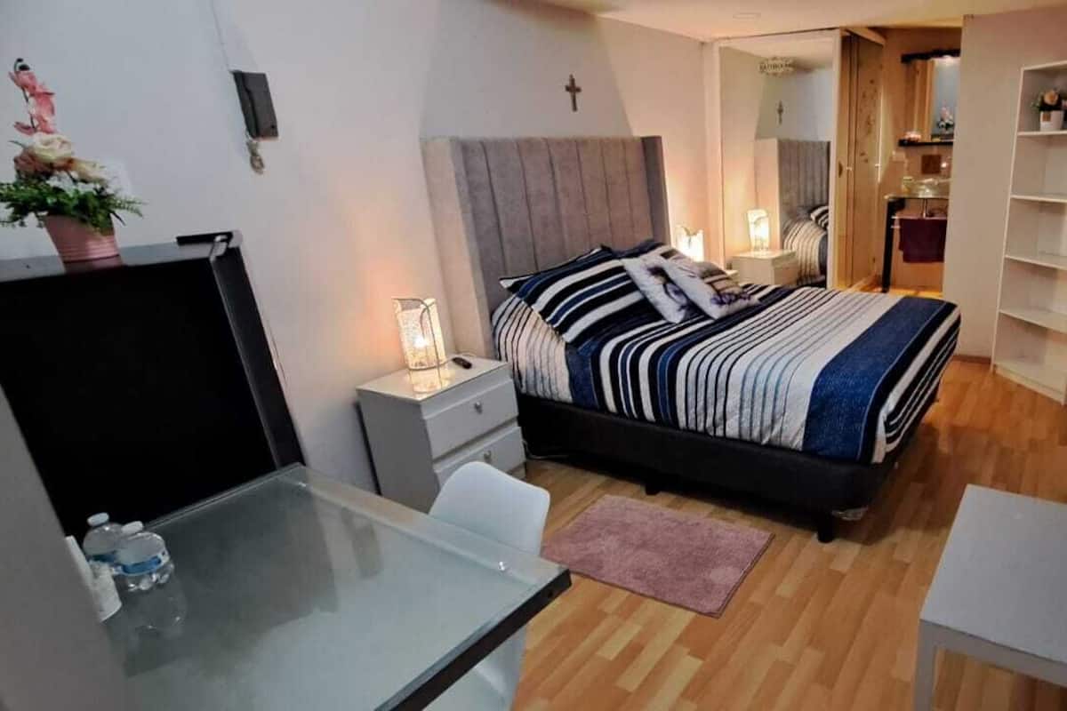 A compact bedroom with a bed, bedside table, lamp, and a small desk with a chair.