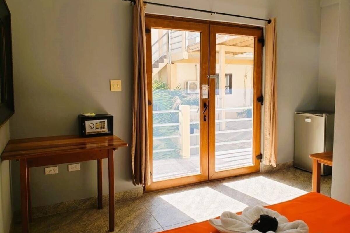 Una habitación con una puerta corrediza de vidrio que da a un balcón, una mesita con una radio, una cama con una colcha naranja y un refrigerador.