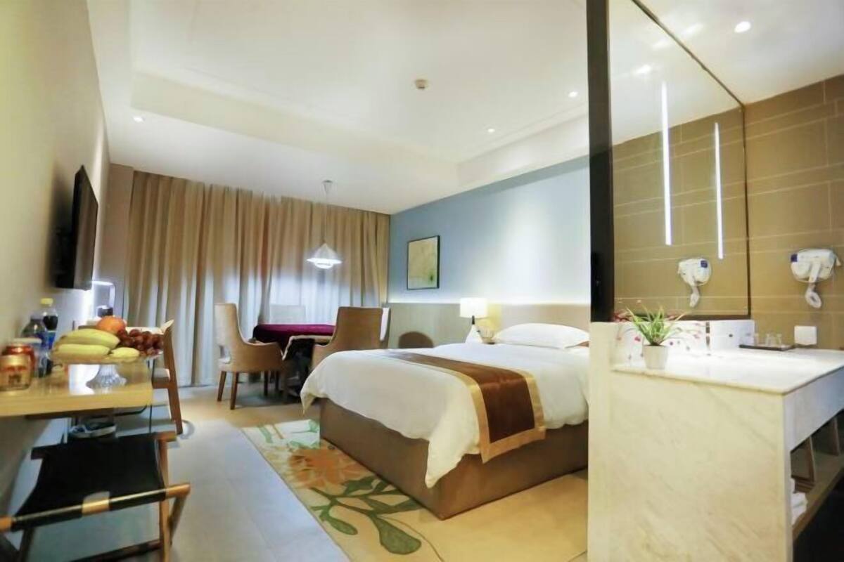 A hotel room with a bed, a desk with a chair, a dining table with chairs, a bathroom with a mirror and sink, and a window with curtains.