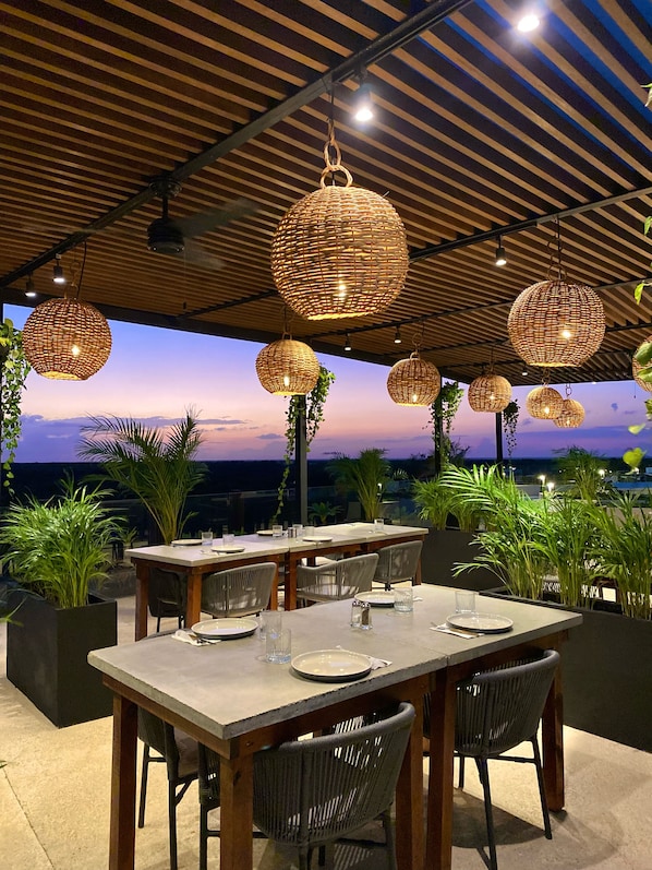 Un área de comedor al aire libre con una mesa dispuesta para cuatro personas, rodeada de sillas de mimbre, bajo un techo de madera con lámparas colgantes esféricas.