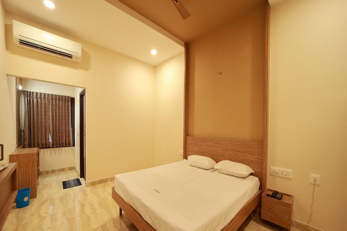 A bedroom with a bed, a ceiling fan, an air conditioning unit, a wooden bedside table, a blue trash bin, and a wall socket.