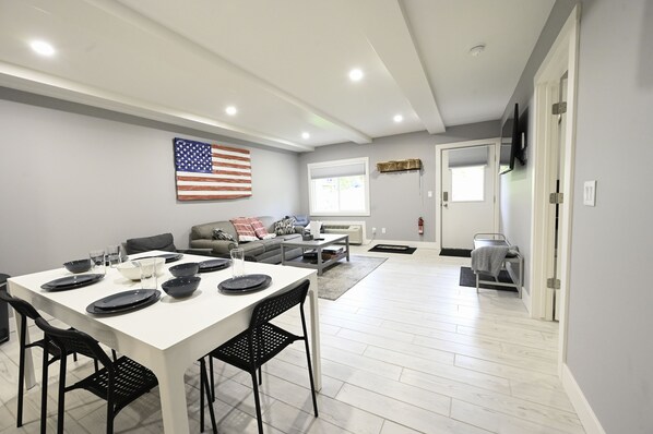 A modern living room with a dining table set for four, a gray sofa, a TV mounted on the wall, and a window with blinds. - Schuss Mountain. 2 bedroom, 1.5 bath Sleeps 6. Pool Dog Friendly boat parking (Mancelona)