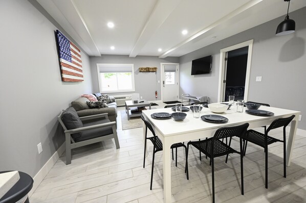 A modern living room with a white dining table set for four, black chairs, a gray sofa, and a large American flag on the wall. - Schuss Mountain. 2 bedroom, 1.5 bath Sleeps 6. Pool Dog Friendly boat parking (Mancelona)