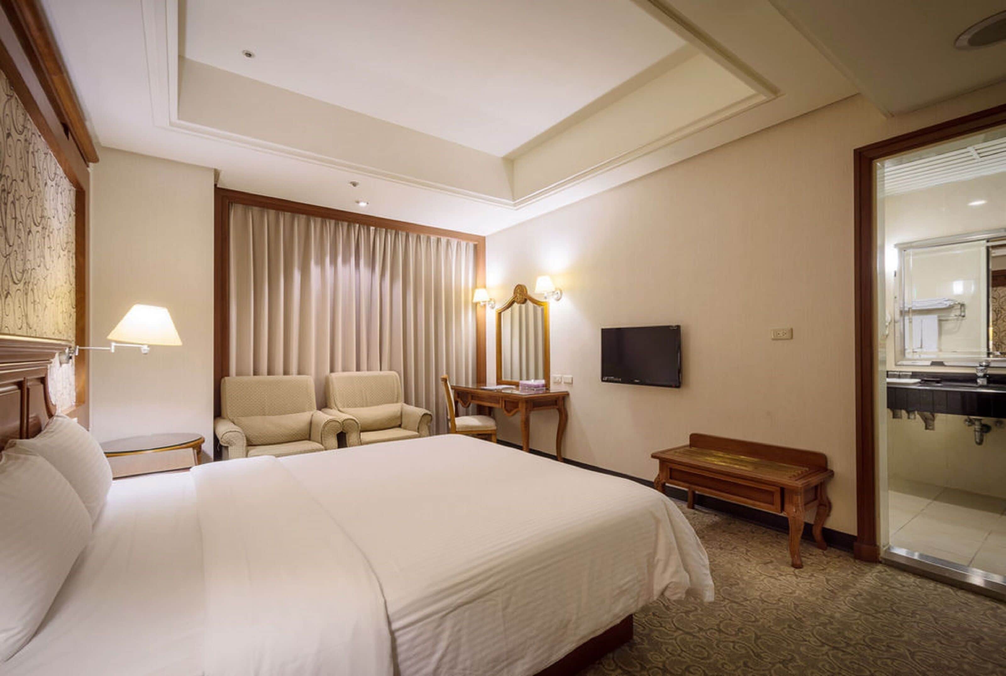 A hotel room with a large bed, a desk, two chairs, a television, and a bathroom visible through an open door.