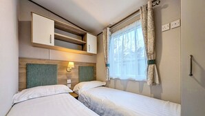 A bedroom with two beds, a wooden headboard, a wall-mounted shelf, a window with curtains, and a wall socket. - 3 bedroom Gold Caravan Pet Friendly (Eamont Bridge)