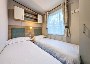 A bedroom with two beds, a wooden headboard, a wall-mounted shelf, a window with curtains, and a wall socket.