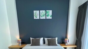 A neatly made bed with white linens and rolled towels, two bedside tables with lamps, a dark blue accent wall, and three framed botanical prints. - Blue Moon Boutique Resort (Sichon)