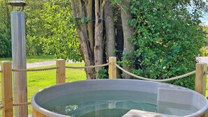 A wooden hot tub with a rope barrier, surrounded by greenery and a tree. - Imagine the fun of camping but with all the luxury comforts of a lodge holiday. (Hale)