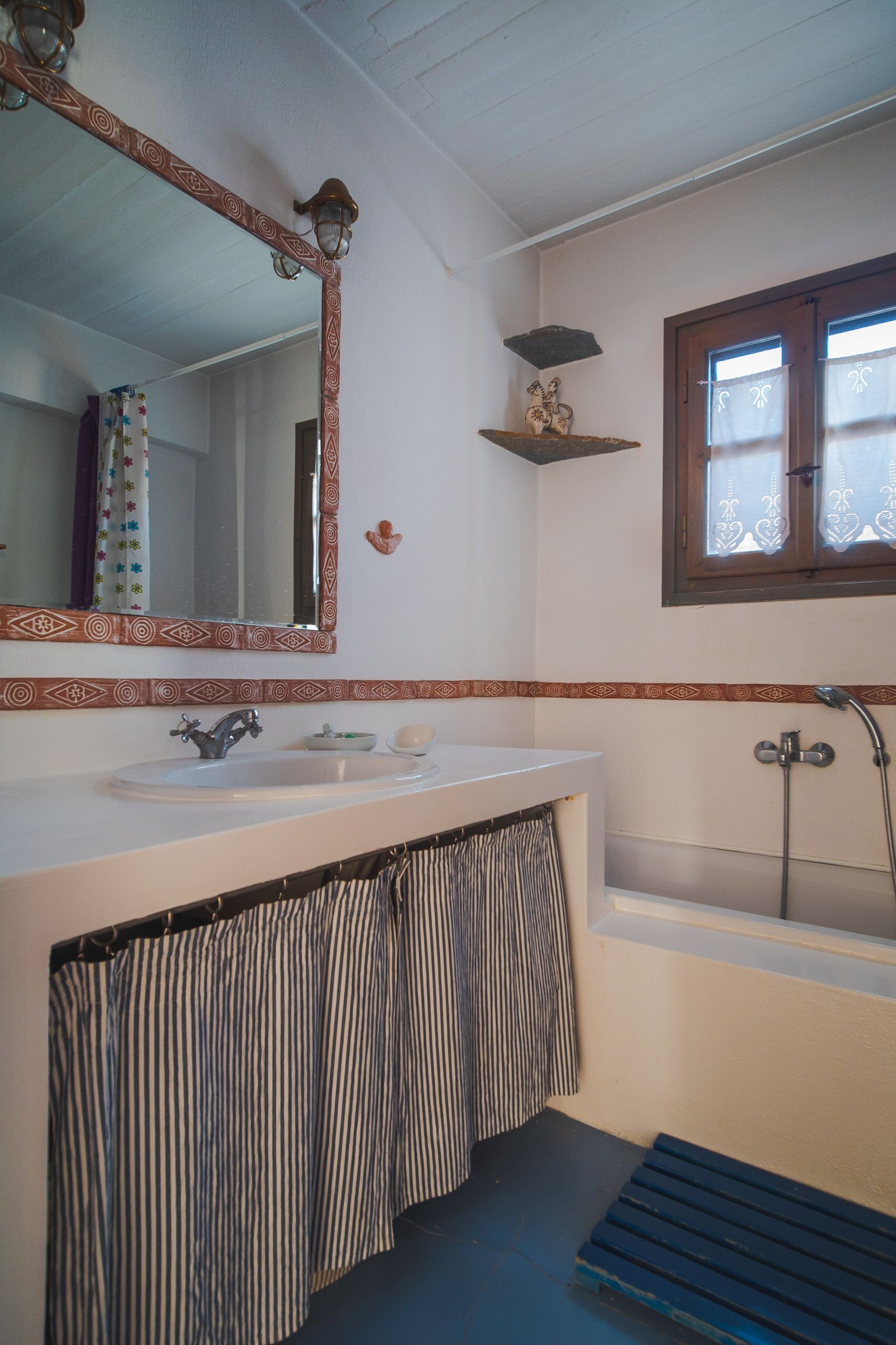 Une salle de bain avec un lavabo blanc, un miroir doté d’un cadre décoratif, une douche et une fenêtre avec des rideaux.