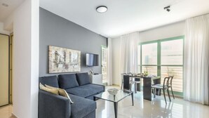 A modern living room with a grey sofa, a glass coffee table, and a dining area with a table and chairs. - MAMAD - Newly Renovated Apartment in Tel Aviv (Tel Aviv)