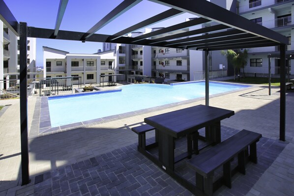 Une piscine dotée d’une pergola et de tables de pique-nique, entourée d’immeubles à plusieurs étages.