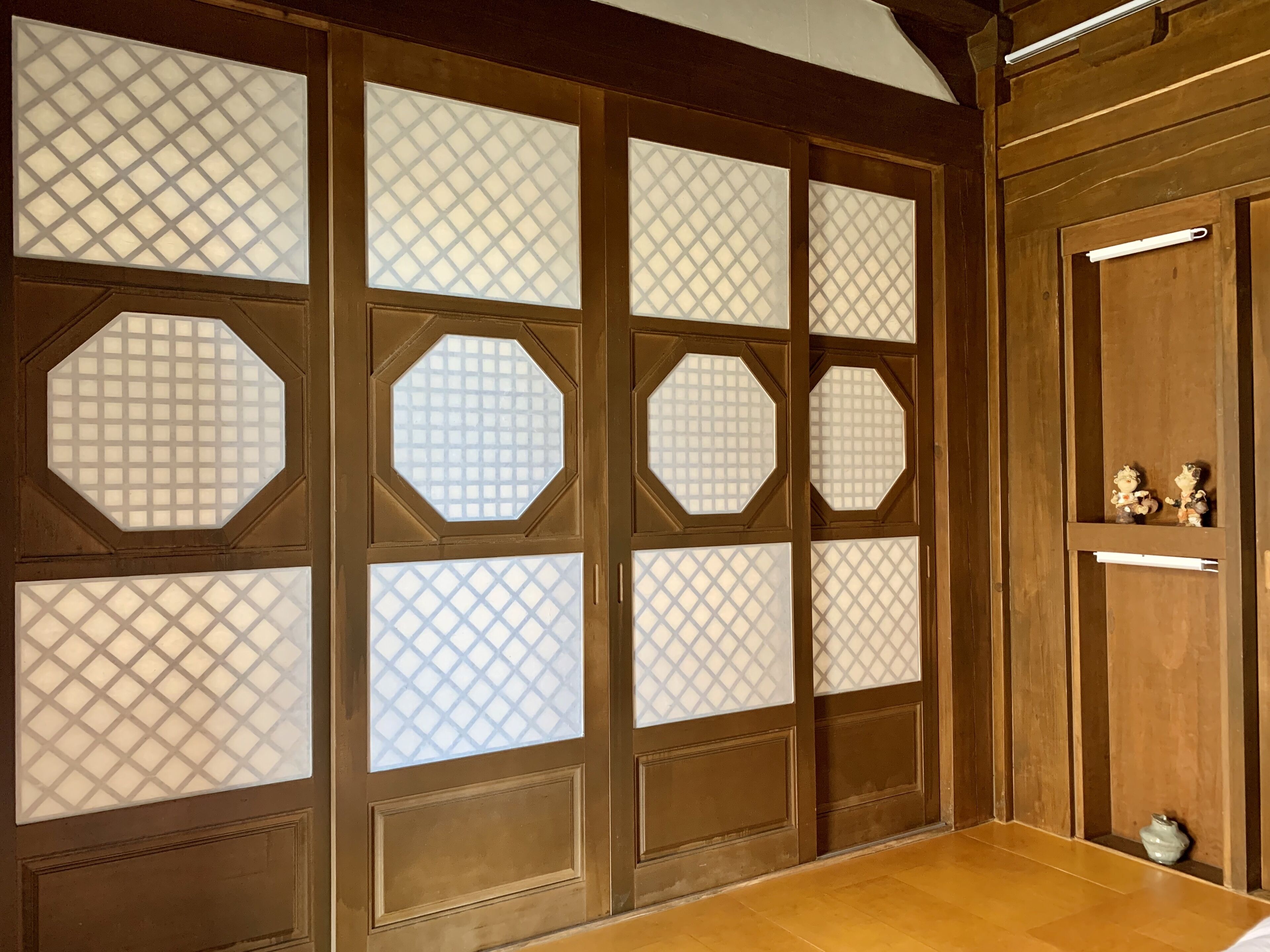A traditional Japanese room with tatami flooring, sliding doors with geometric patterns, and a shelf with decorative items.