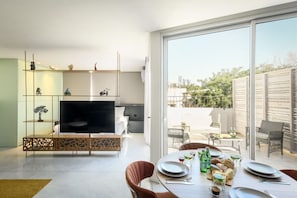 A modern living room with a flat-screen TV, a dining area with a table set for two, and a large sliding glass door leading to an outdoor patio.