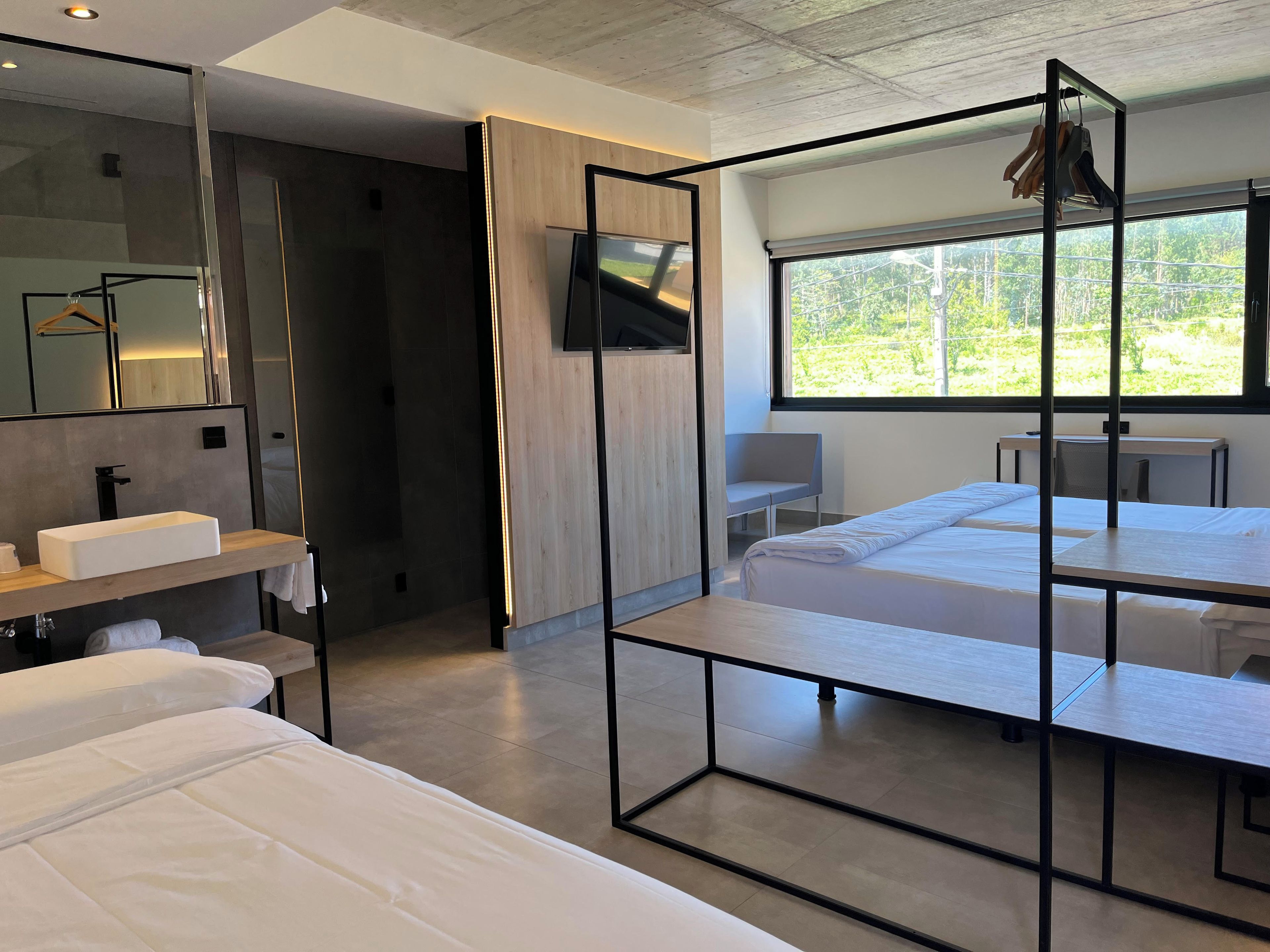 A modern hotel room with a large bed, a bedside table, a mirror, a TV, and a window with a view of greenery.