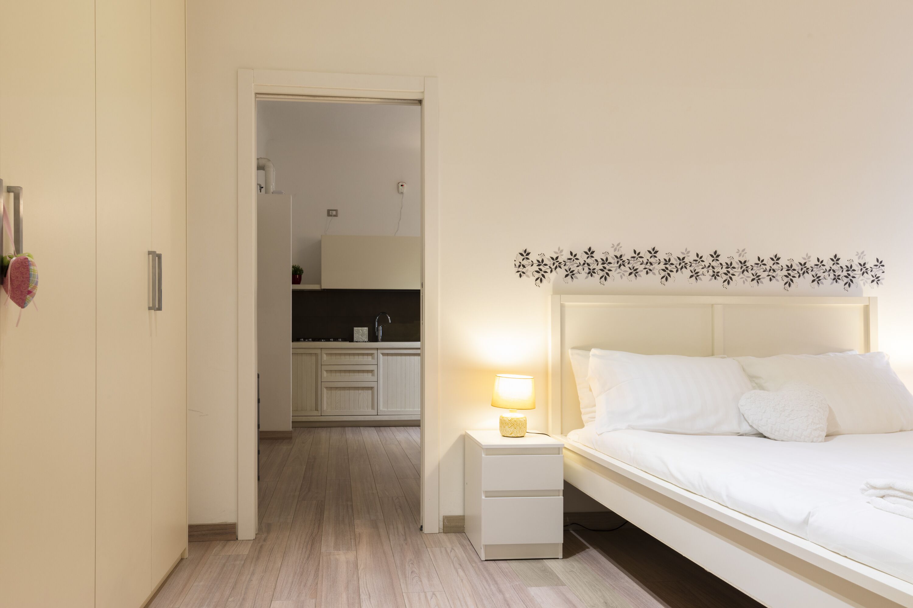 Un dormitorio con cama, mesita de noche y una mesilla con lámpara. Se ve una puerta que da a un baño y una zona de cocina al fondo.
