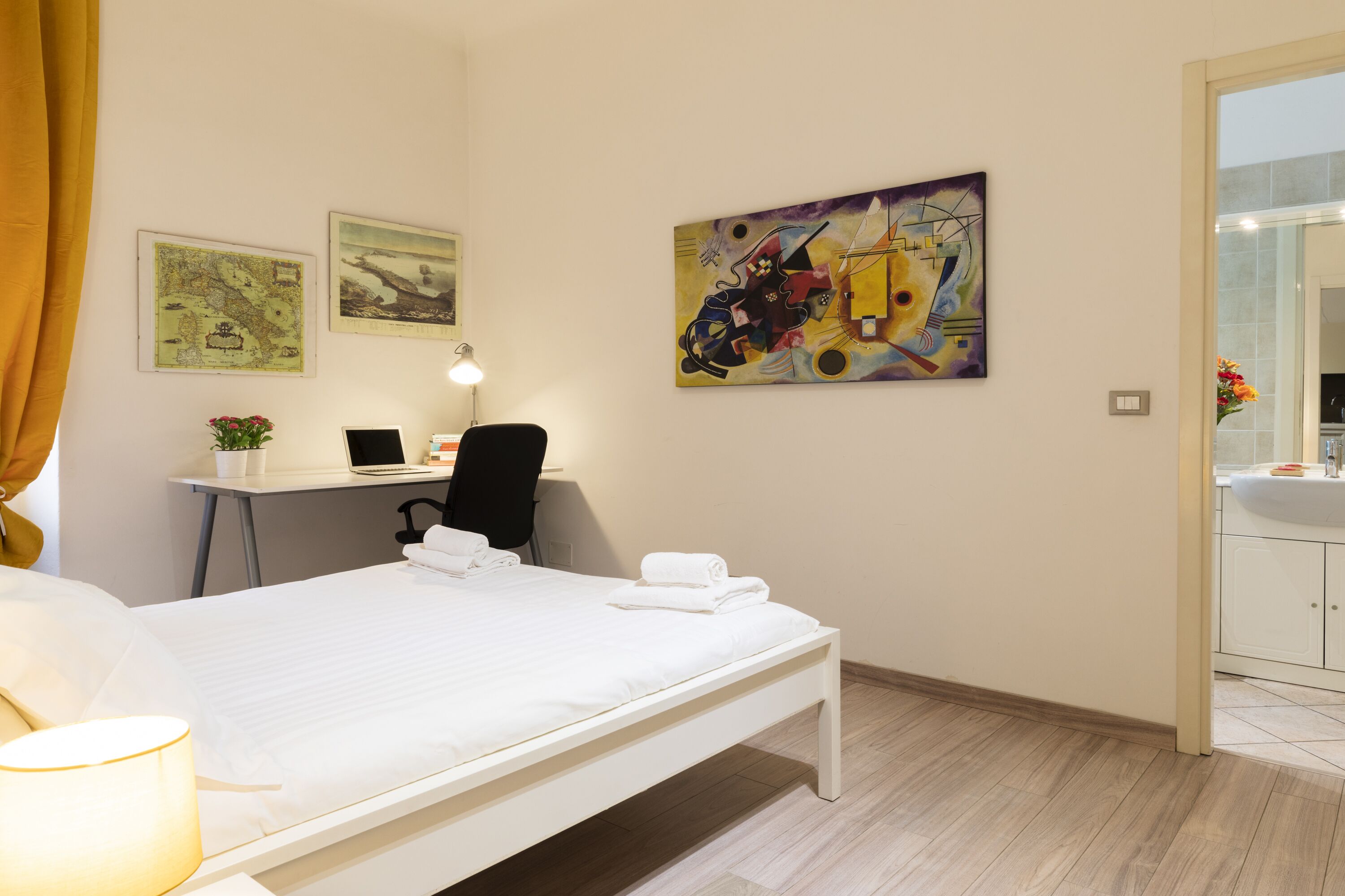 A hotel room with a bed, desk, chair, and a painting on the wall.