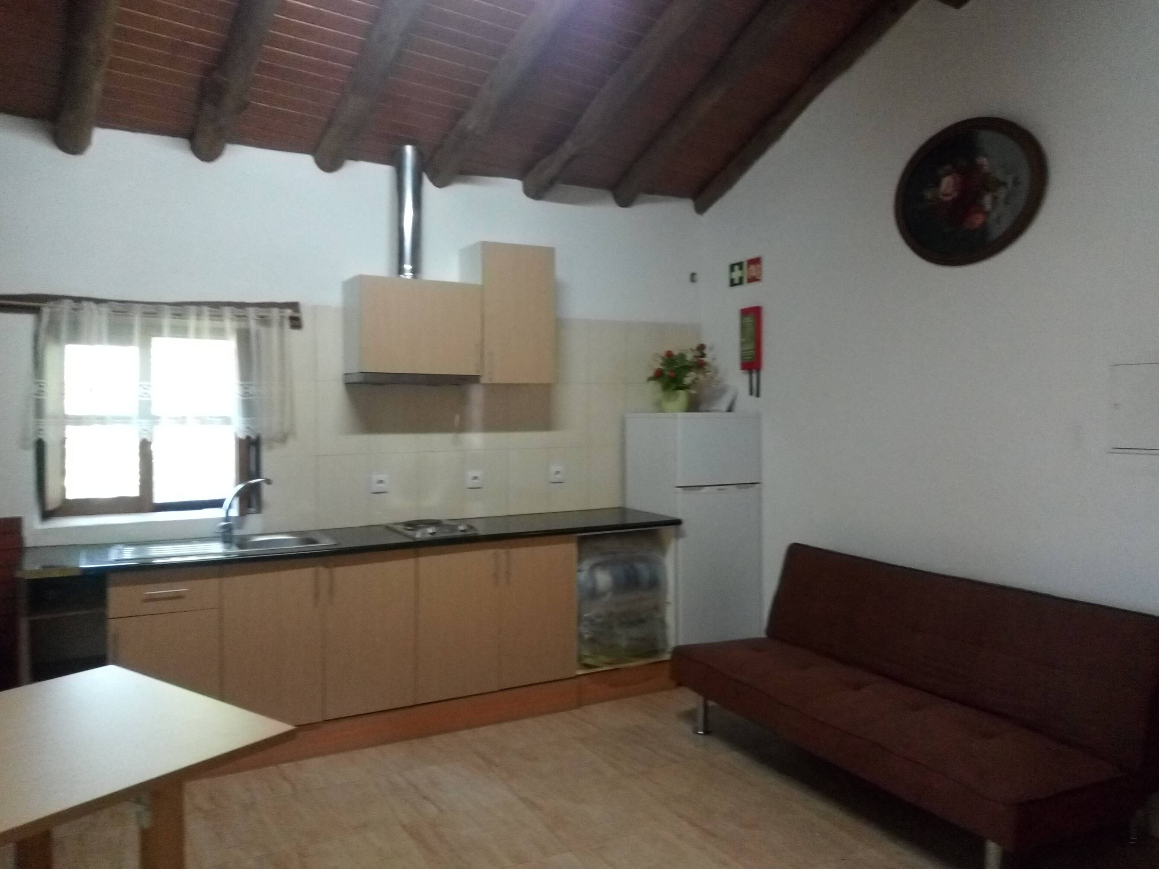 Una cocina compacta con pileta, estufa y alacenas. Se observa un sofá rojo y una mesita.