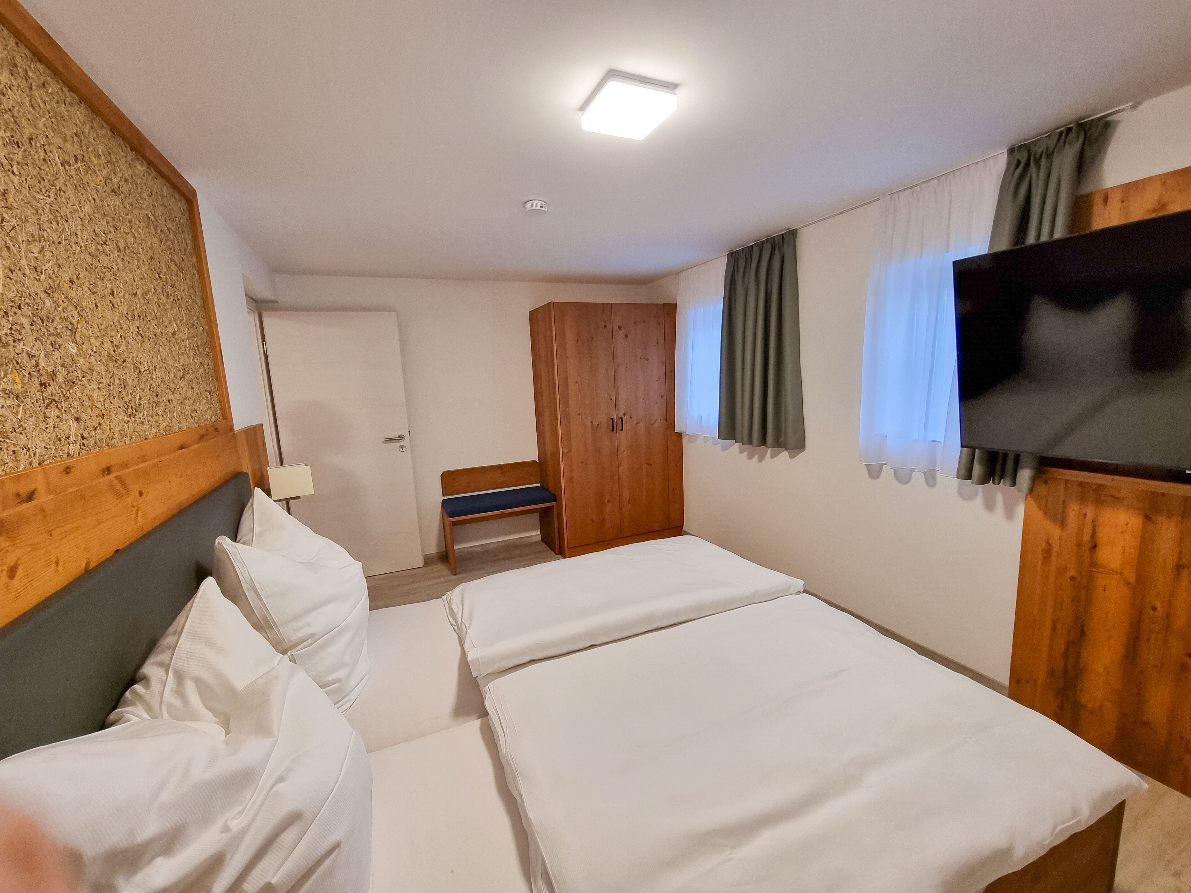 Habitació d'hotel amb dos llits, un armari de fusta, una panxa, una televisió i una làmpada al sostre.