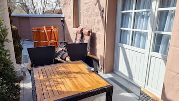 Une terrasse extérieure avec une table en bois et des chaises en osier, une porte-fenêtre coulissante et un petit arbre.
