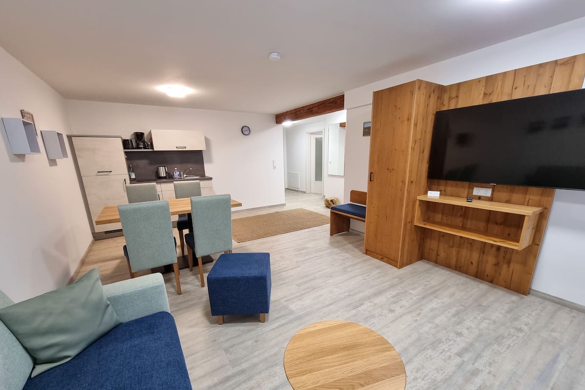 A modern living room with a sofa, ottoman, and round coffee table. There is a TV mounted on a wooden panel, a kitchen area with a dining table and chairs, and a dog in the background.