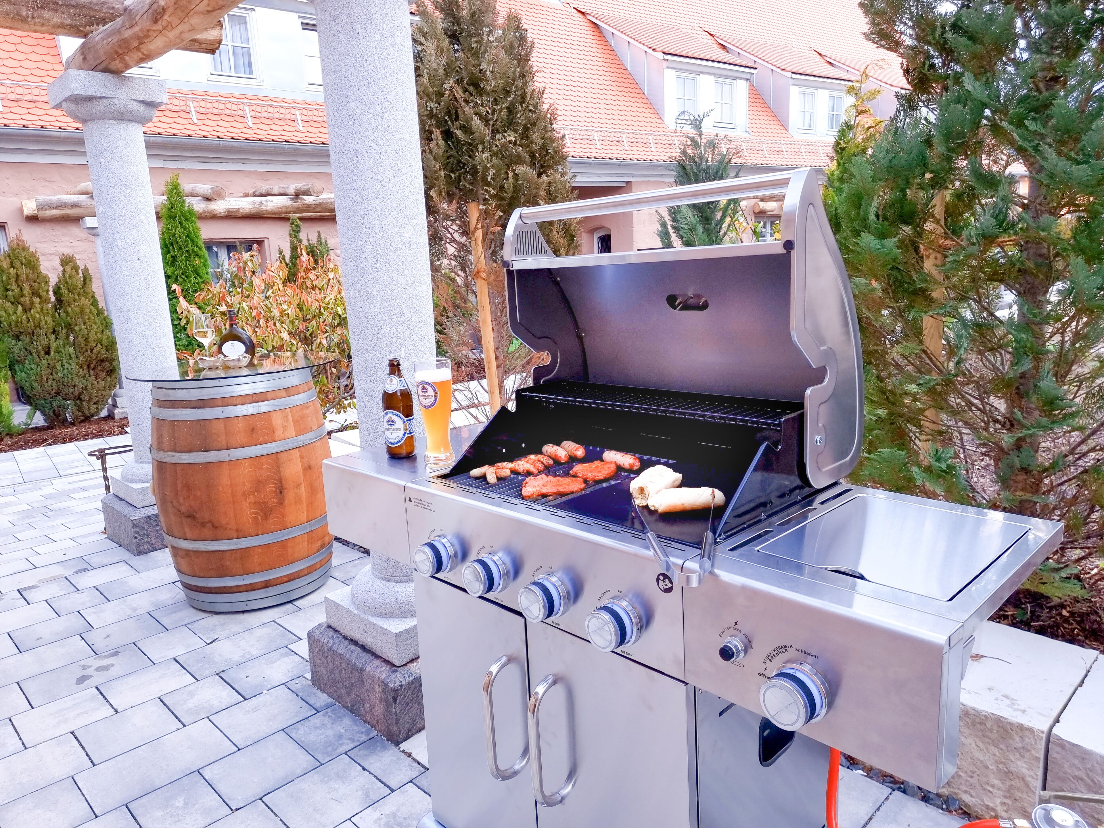 Un barbecue installé à l’extérieur, avec des saucisses et une bouteille de bière, le tout sur fond d’une maison et d’arbres.