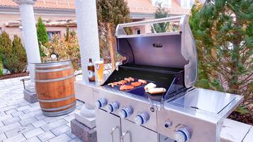 Un barbecue installé à l’extérieur, avec des saucisses et une bouteille de bière, le tout sur fond d’une maison et d’arbres.