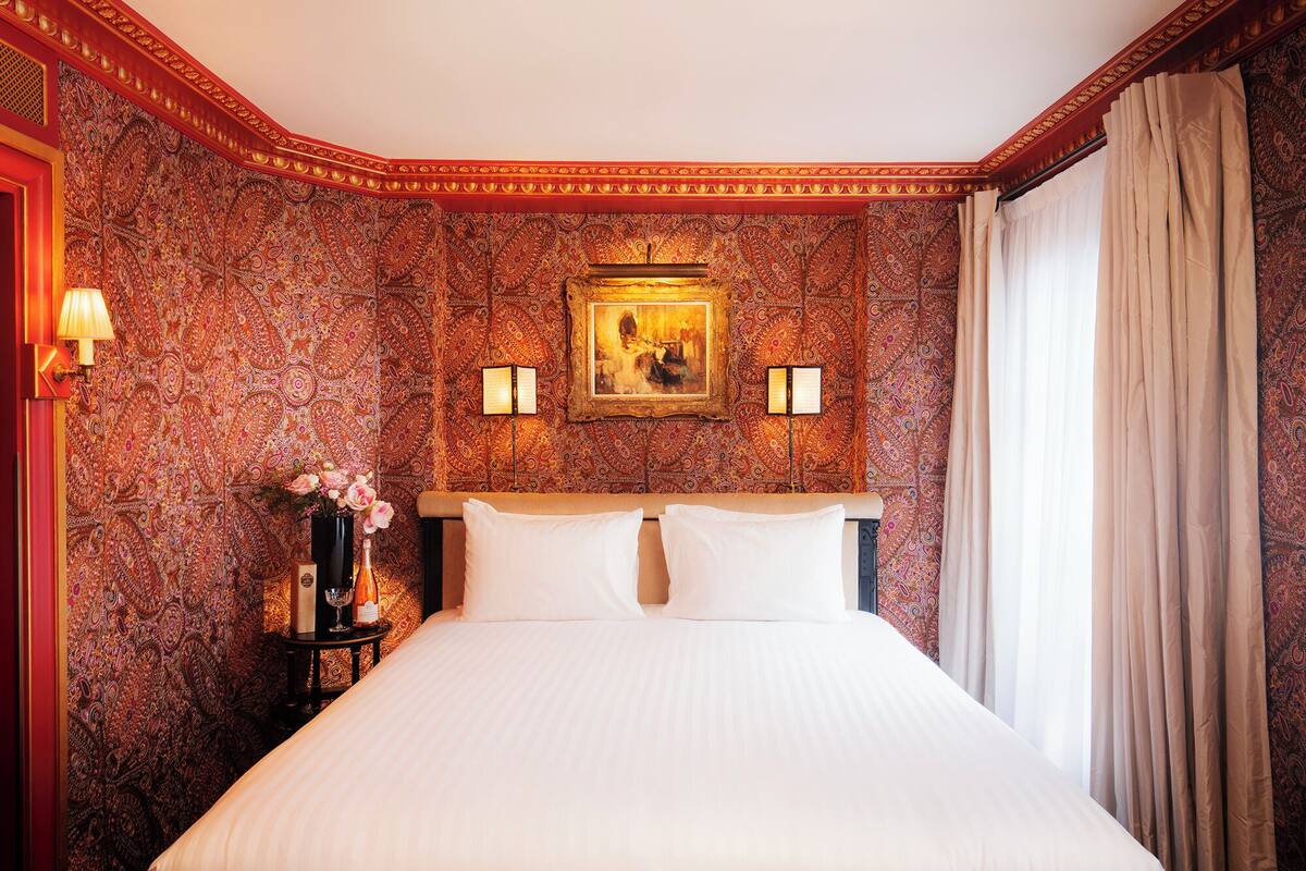 A hotel room with a large bed, two bedside lamps, a painting above the bed, and a patterned wallpaper.