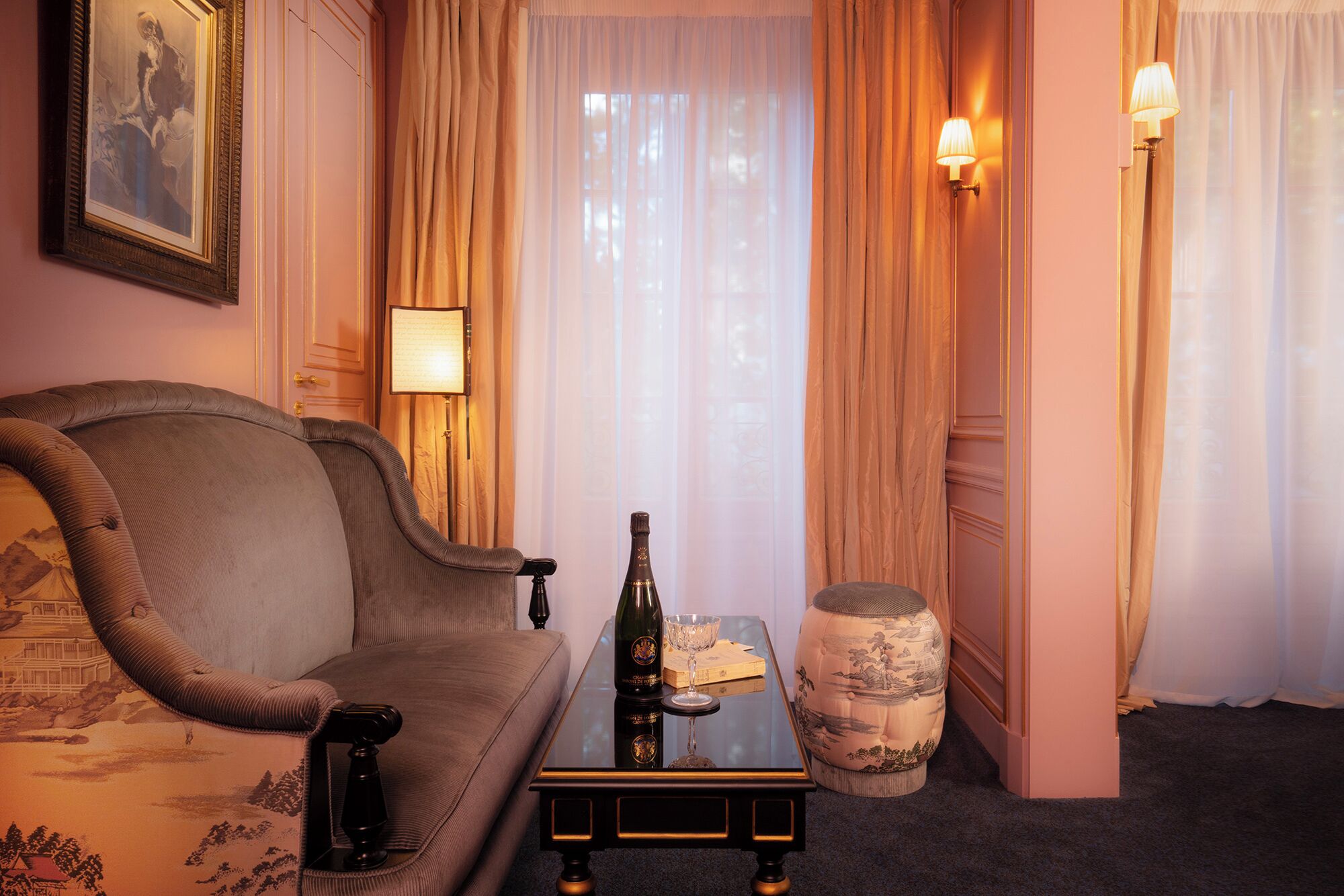 A hotel room with a sofa, a small table with a bottle and glass, a decorative vase, and a framed picture on the wall.