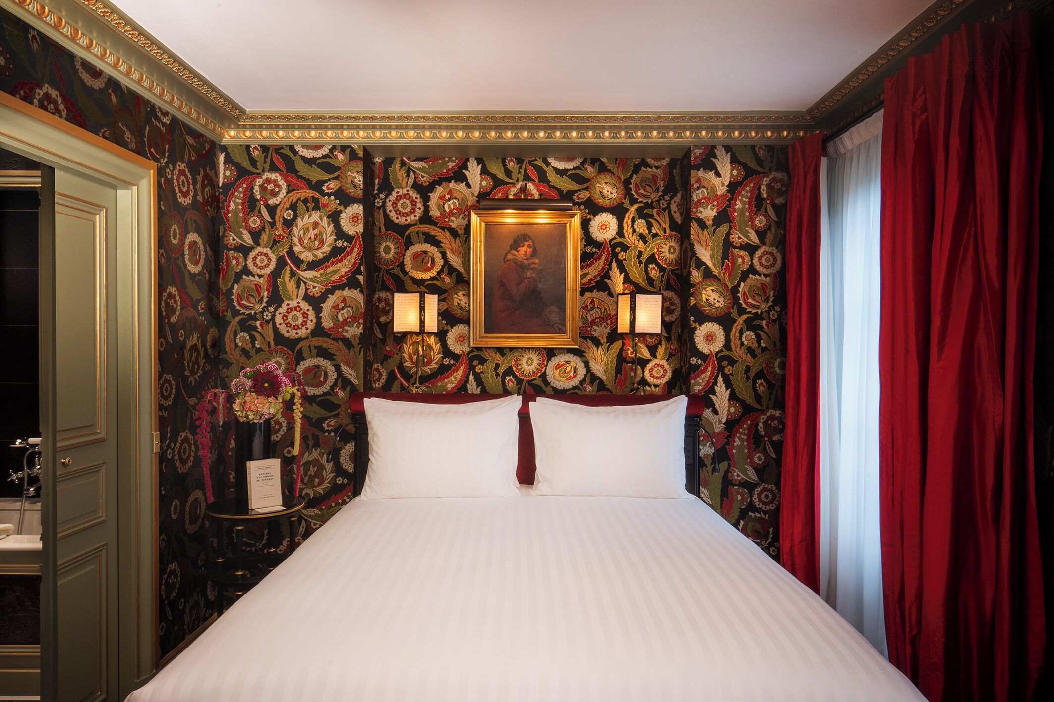 Quarto de hotel com cama, papel de parede floral, quadro emoldurado, duas arandelas e cortinas vermelhas.