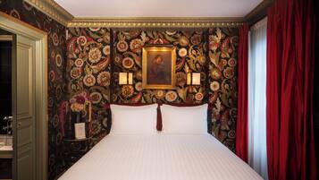 Quarto de hotel com cama, papel de parede floral, quadro emoldurado, duas arandelas e cortinas vermelhas.
