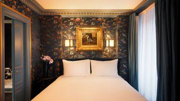 Een hotelkamer met een groot bed, bloemenbehang, een ingelijst schilderij, twee wandlampen en een raam met gordijnen.