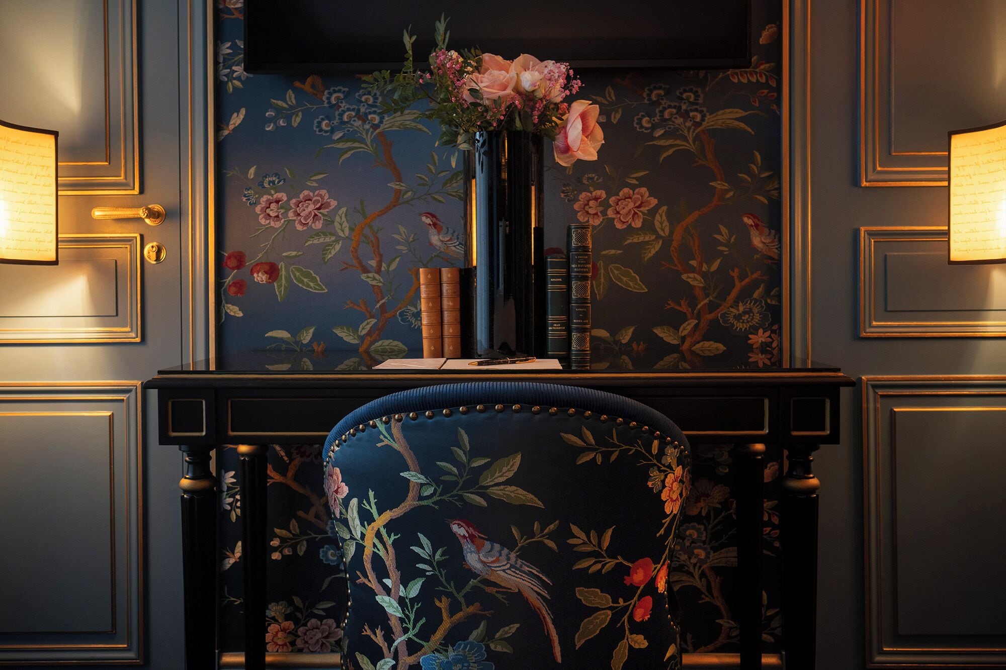 A room with a dark blue floral wallpaper, a black bookshelf with books, a vase with flowers, and a chair with embroidered birds.