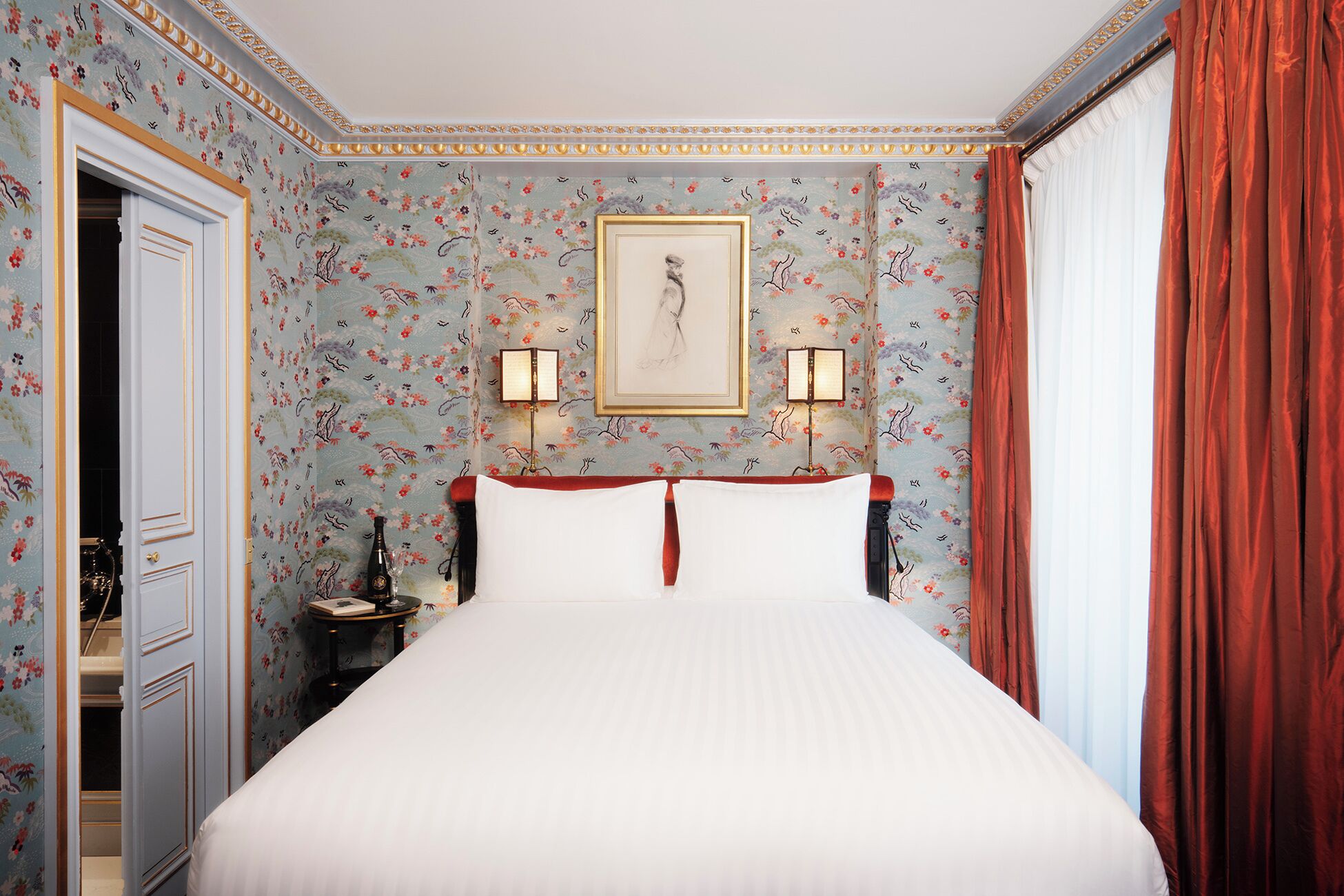 Habitació d'hotel amb paper pintat floral, un llit amb roba de llit blanca, dues làmpades penjades a la paret i una obra d'art emmarcada sobre el llit.
