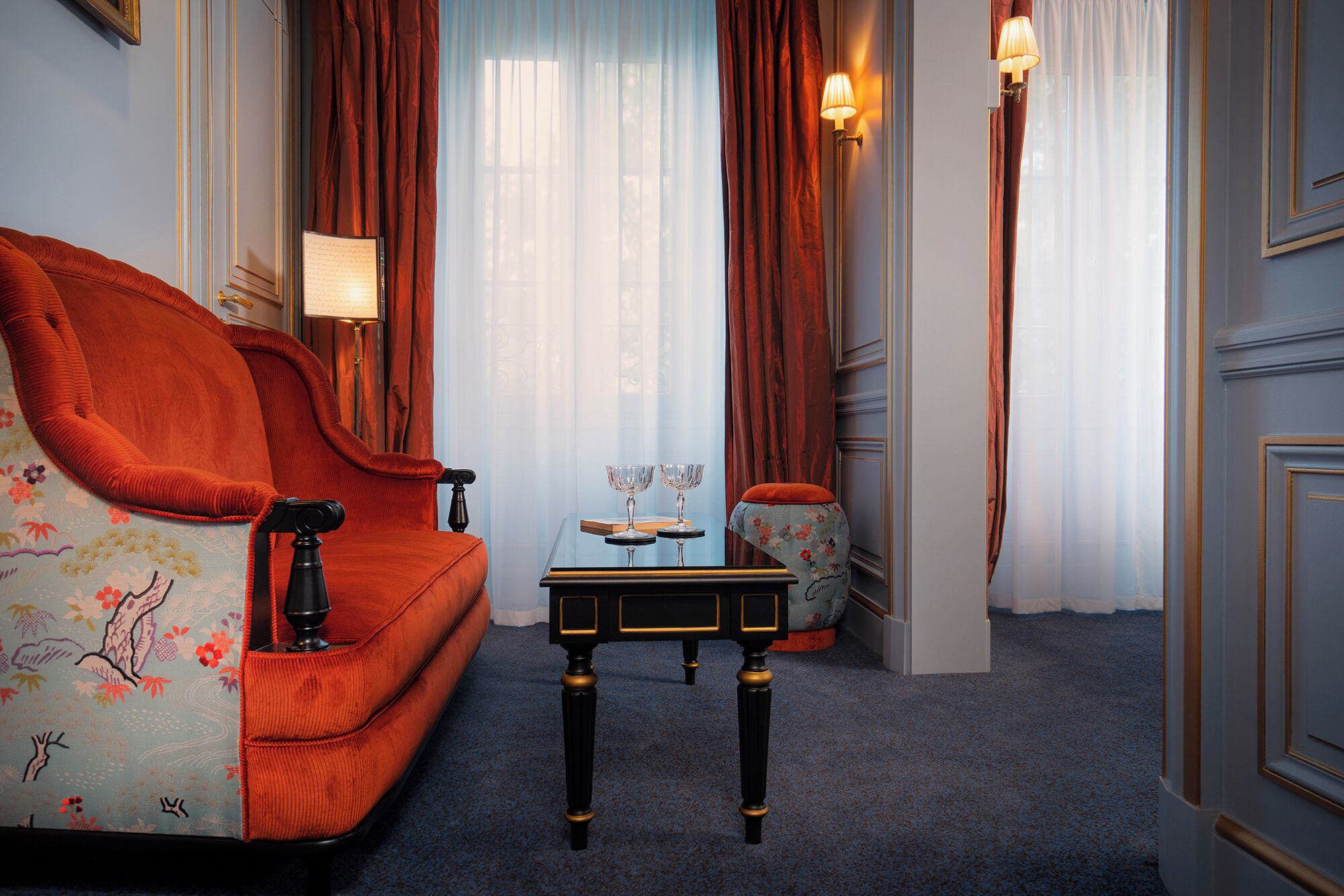 A room with a red sofa, a small table with two wine glasses, and a lamp.