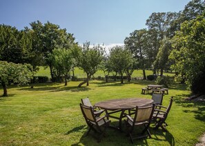Une table ronde en bois, entourée de quatre chaises, se trouve dans un espace extérieur verdoyant, bordé d’arbres.