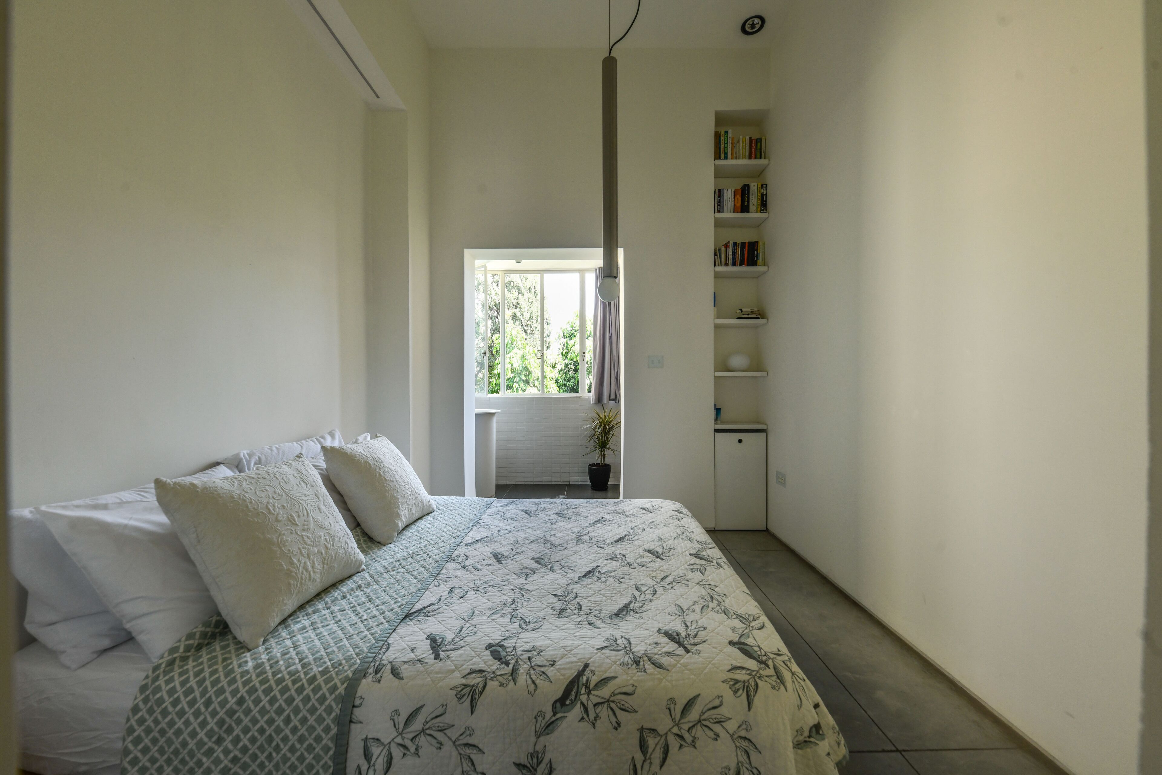 A bedroom with a bed, pillows, a bedside table, a window, a bookshelf, and a potted plant.