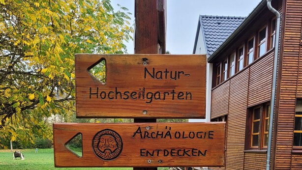 A wooden signpost with multiple directional arrows and labels for Naturhochseilgarten, Archäologie Entdecken, and Archäologischer Landschaftspark. - Cozy house with panoramic views (Nettersheim)