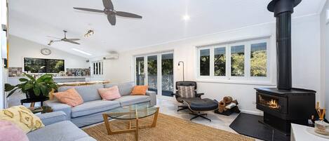 Ein geräumiges Wohnzimmer mit einer Couch, Couchtisch und Kamin. Deckenventilatoren und ein großes Fenster mit Blick auf Bäume.